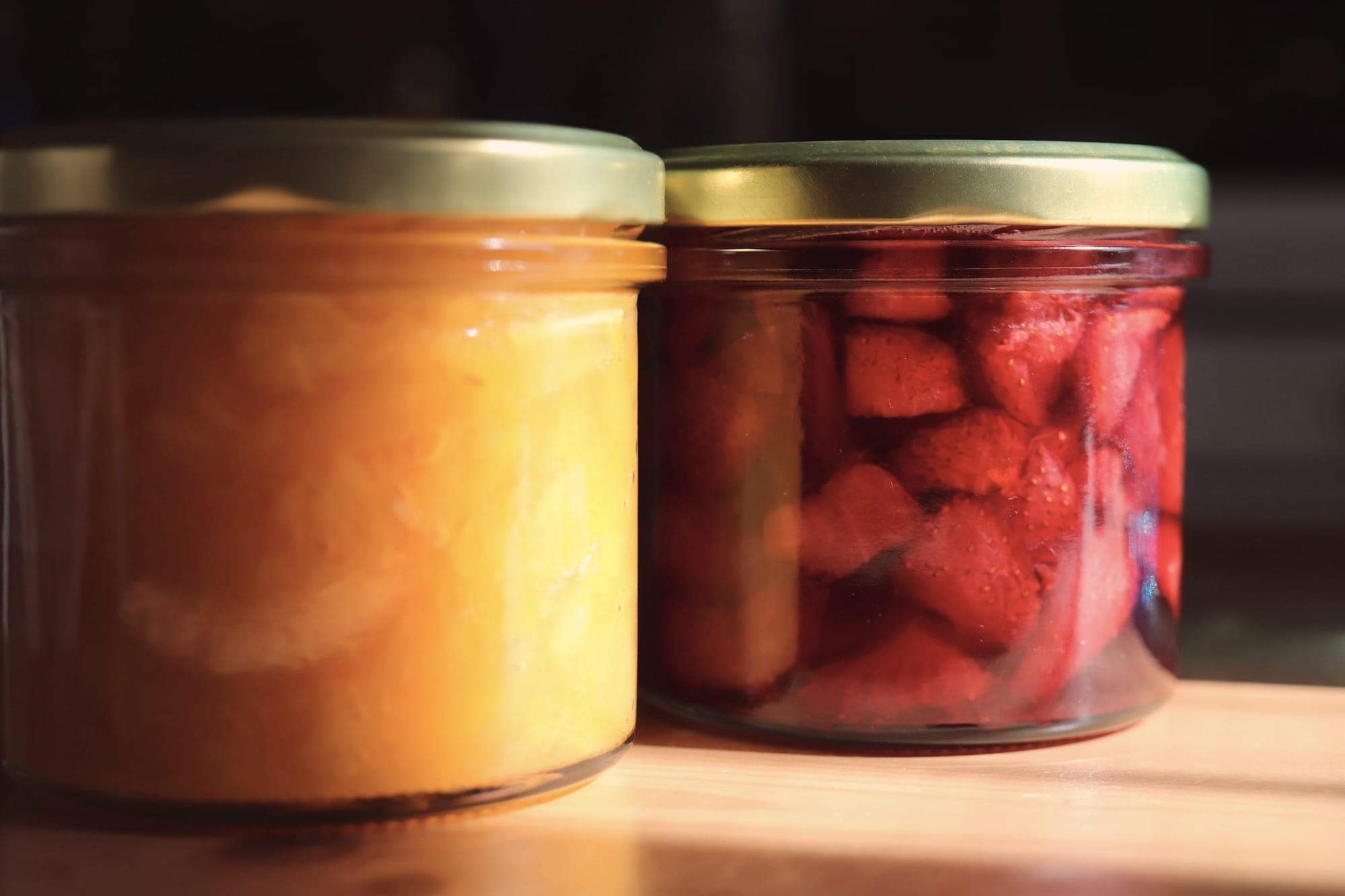 Deux bocaux en verre posés au soleil sur une planche, l’un rempli de fruits jaunes, l’autre de fraises.