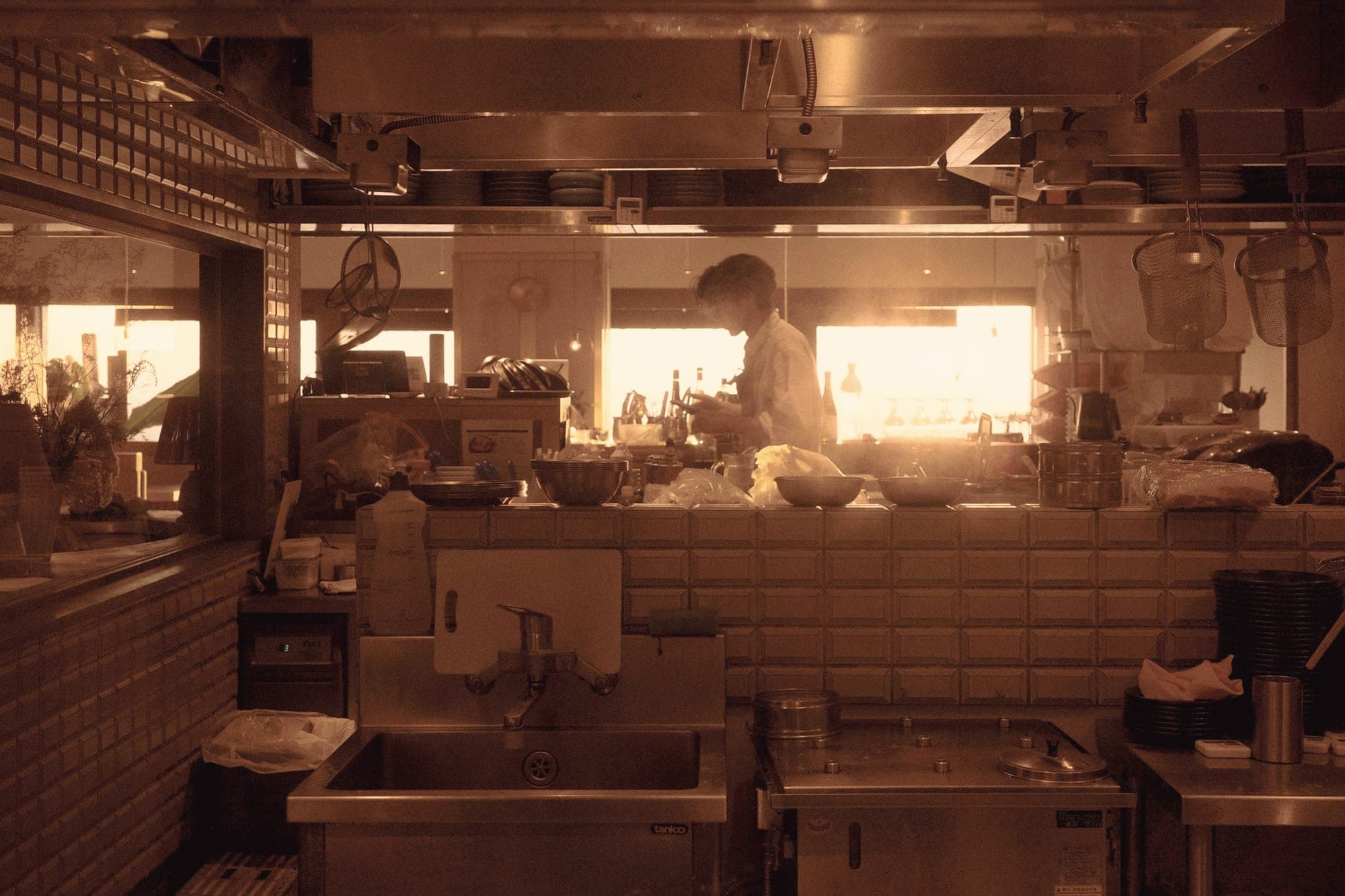 La cuisine ouverte vue depuis la salle, à contre-jour, avec un cuisinier au travail devant les fenêtres.