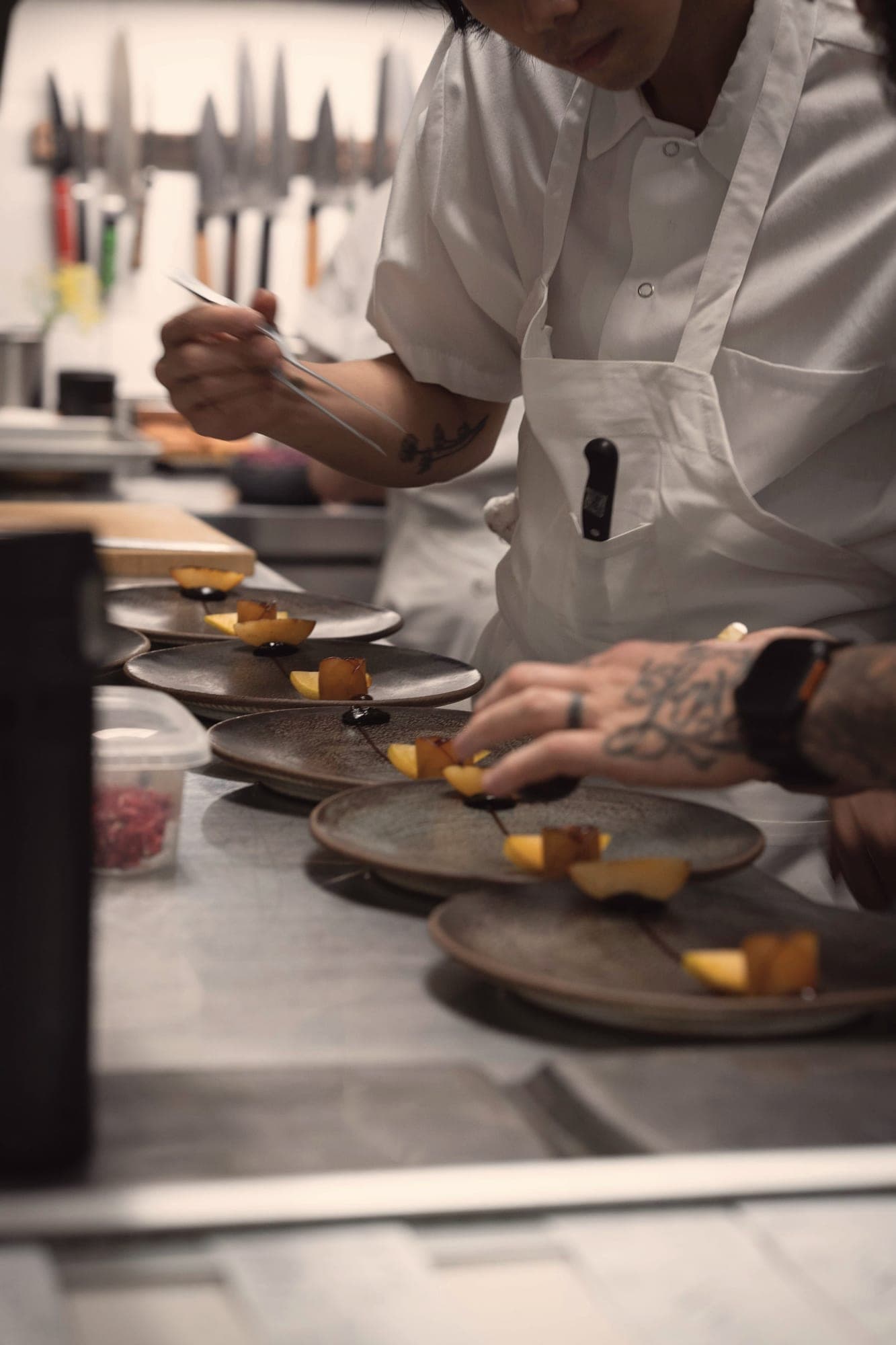 Deux cuisiniers dressent à la pince des morceaux de courge rôtie sur une rangée d’assiettes en grès.