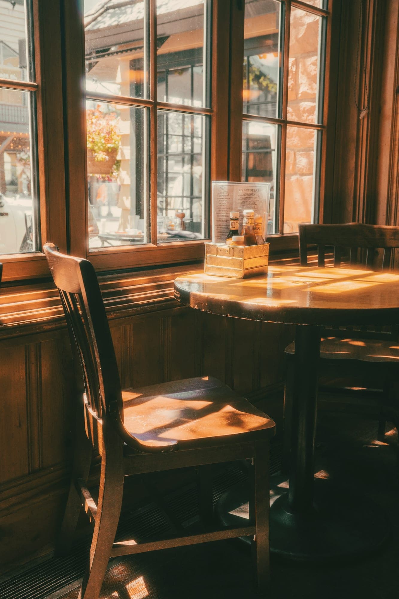 Une table ronde en bois et sa chaise, baignées de soleil près d’une grande fenêtre à petits carreaux.