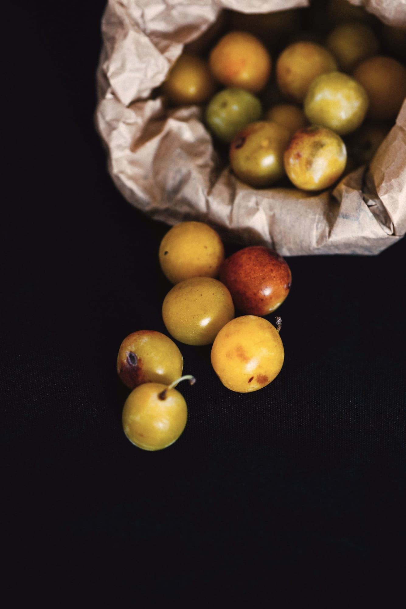 Des mirabelles dorées, tachetées de rouge, s’échappent d’un sac en papier kraft sur un fond sombre.