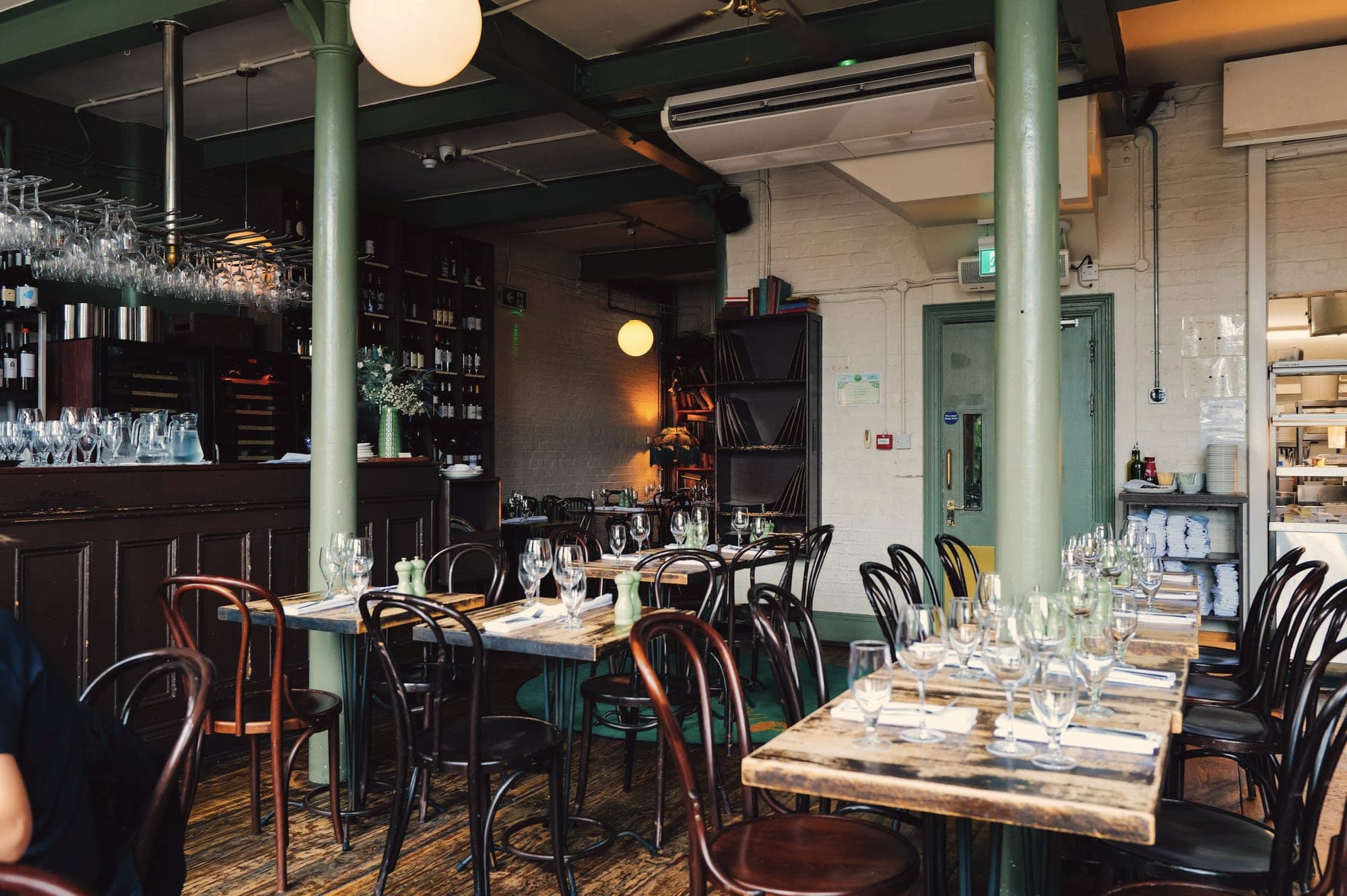 La salle du bistrot : tables en bois dressées, chaises bistrot, colonnes vertes et grand comptoir sombre.